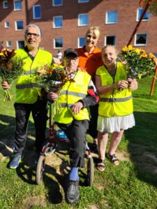 Tre personer i varselväst med blombuketter omgiven av vdn i orange pikétröja